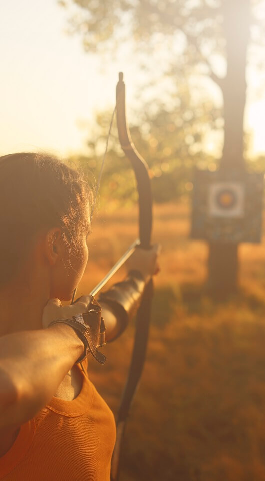 Mujer con un arco apuntando al blanco, se encuentra en un espacio abierto en el bosque
